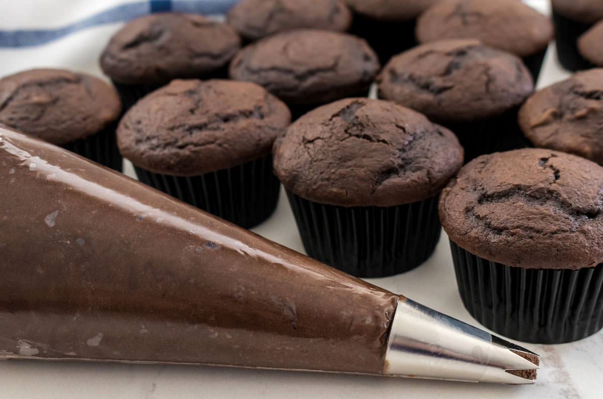 Closeup on a decorating bag filled with the Dark Chocolate Frosting sitting in front of a batch of unfrosted chocolate cupcakes.