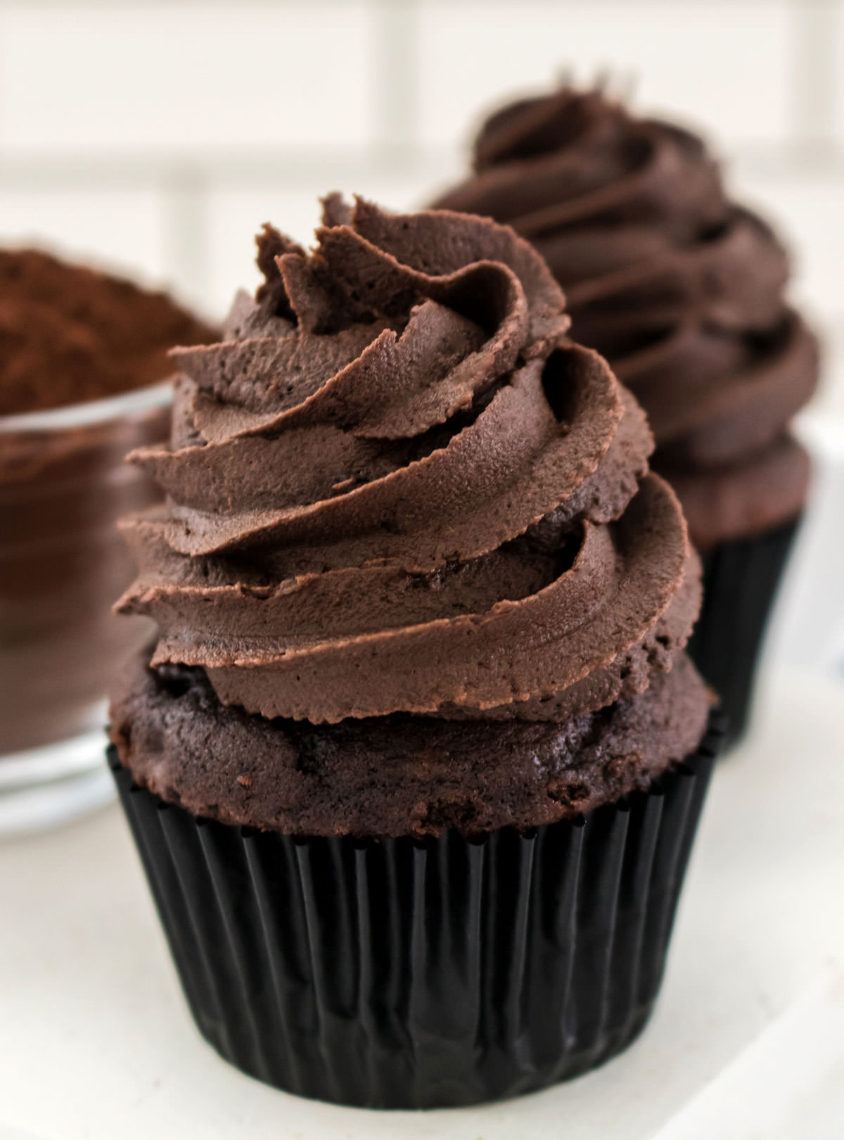 Closeup on a chocolate cupcake topped with The Best Dark Chocolate Buttercream Frosting sitting next to a glass bowl filled with cocoa powder.