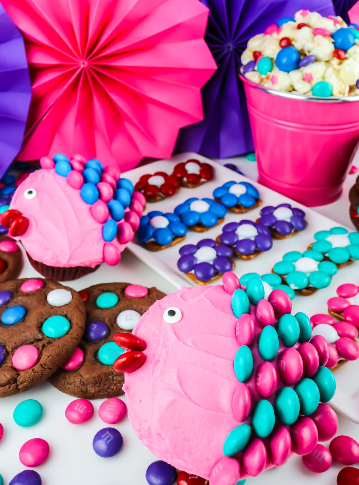 A series of desserts decorated with colored M&M including pretzel bites, popcorn, cupcakes and cookies.