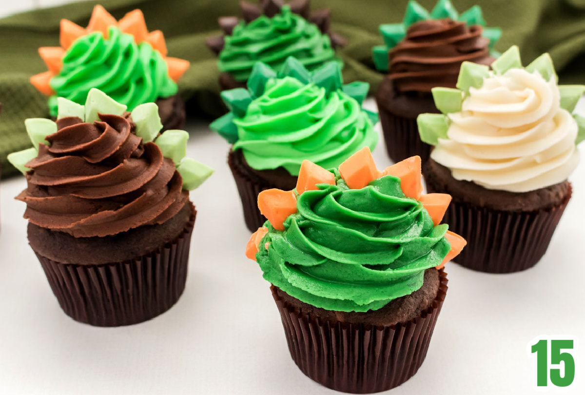 Closeup on seven Dinosaur cupcakes sitting on a white table in front of a green kitchen towel.