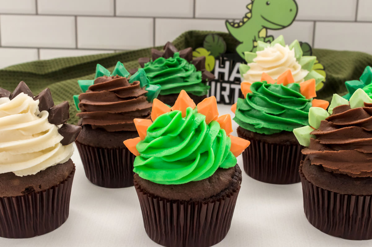 Closeup on eight Dinosaur Cupcakes sitting on a white table with a green kitchen towel and a cartoon dinosaur in the background.
