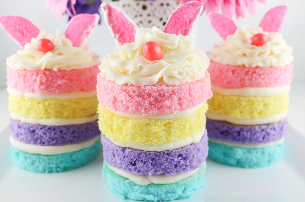 Closeup of three Easter Bunny Mini Cakes sitting on a white surface in front of a white vase filled with pink and purple daisies.