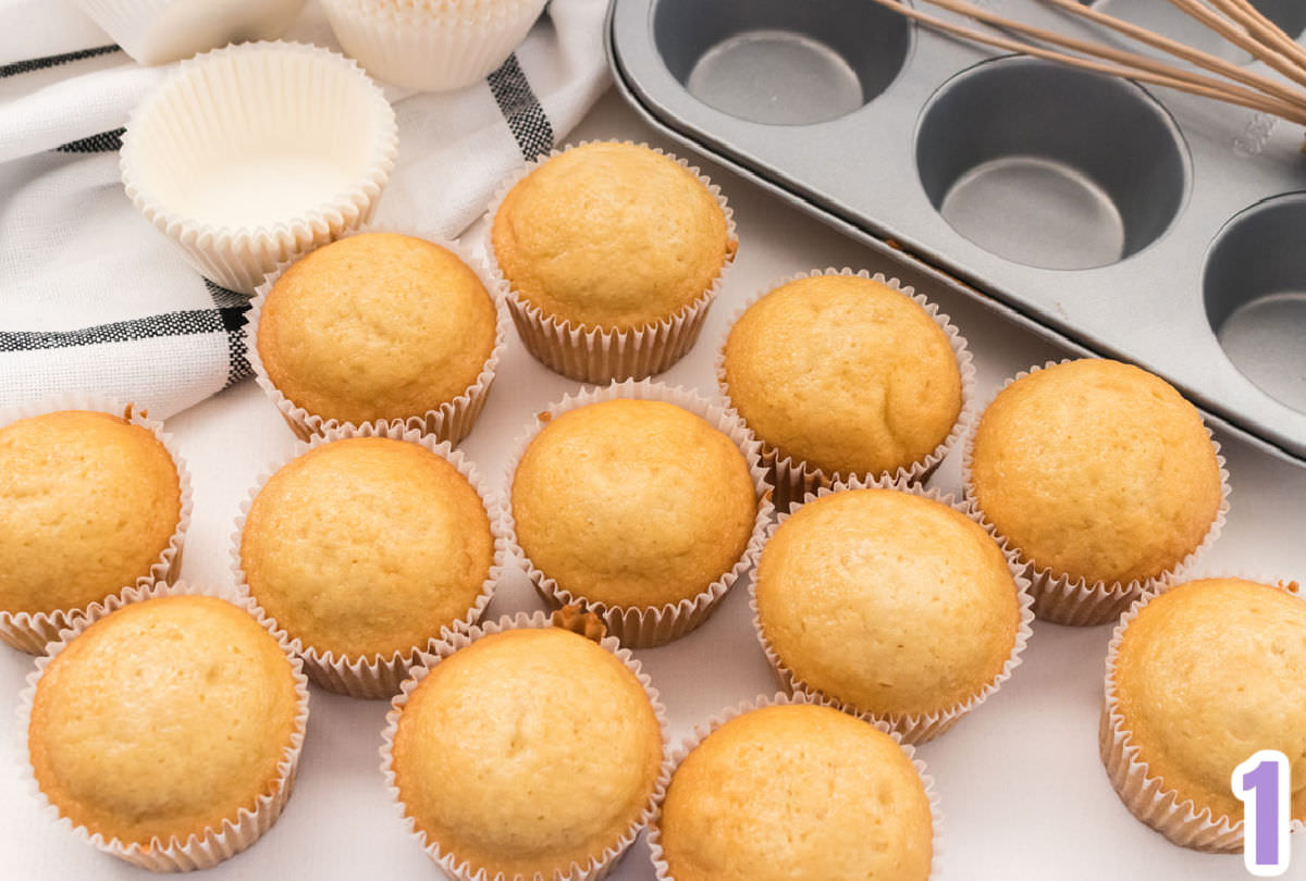 Overhead shot of 12 vanilla cupcakes sitting on a white surface in front of a cupcake tin and a white and black table linen.