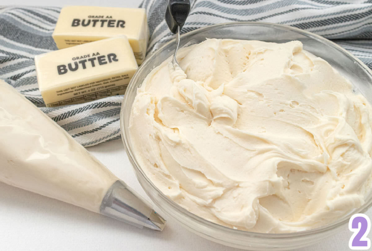 Glass mixing bowl filled with buttercream frosting sitting next to a decorating bag filled with frosting and two sticks of butter.