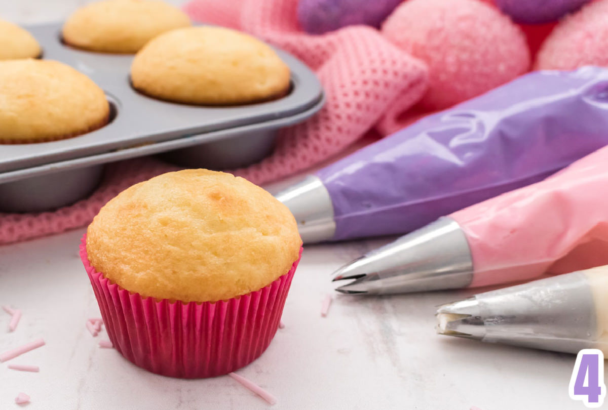 Closeup of a vanilla cupcake in a pink cupcake liner sitting next to frosting decorating bags and a pan full of vanilla cupcakes.