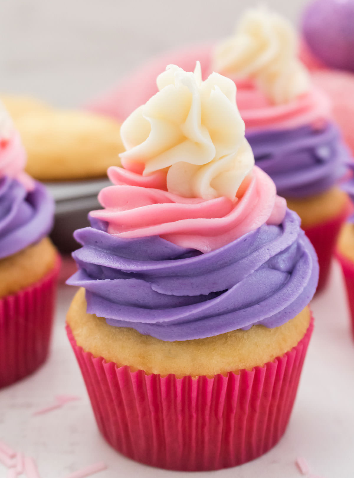 Close up of an Easter Cupcake sitting on a white table in front of more cupcakes, a cupcake tin and a pink table linen.