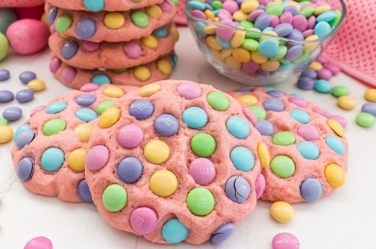 Three Easter M&M Sugar Cookies sitting on a white table surrounded by a bowl of pastel M&M's and another stack of cookies.