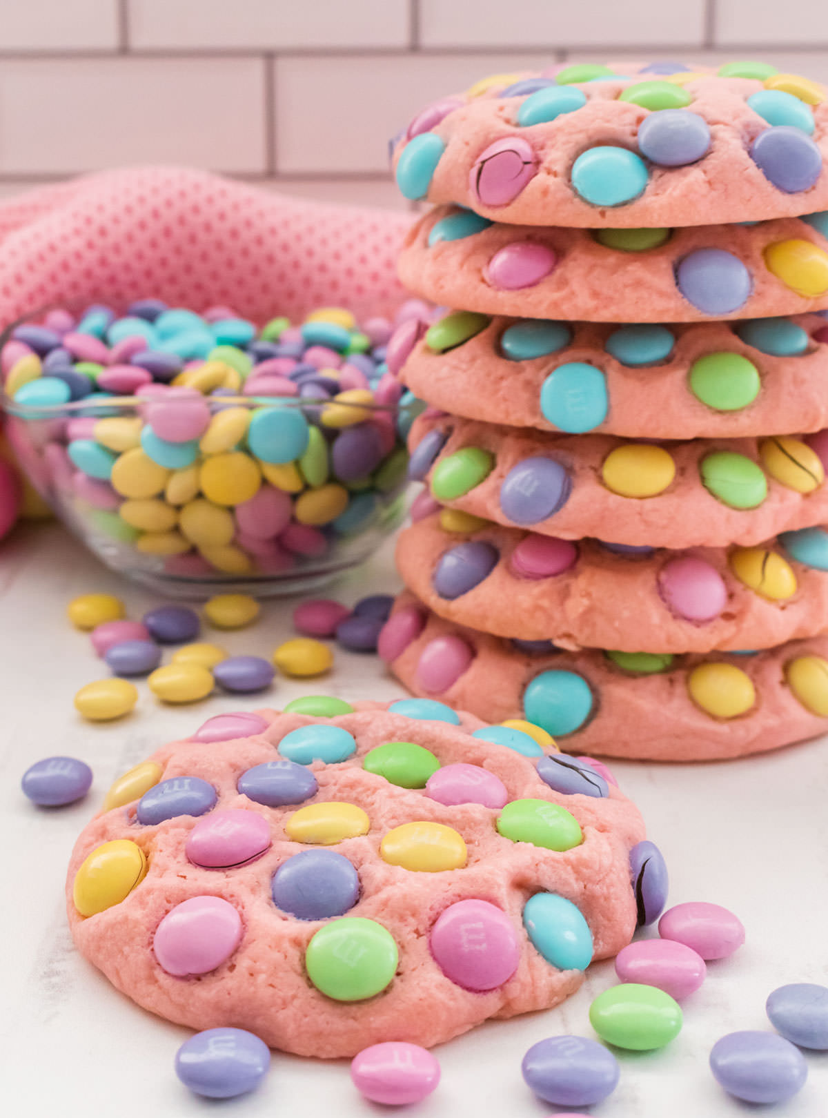 Closeup on an Easter M&M Sugar Cookie sitting on a white surface in front of a larger stack of cookies and a glass bowl filled with pastel M&M's.