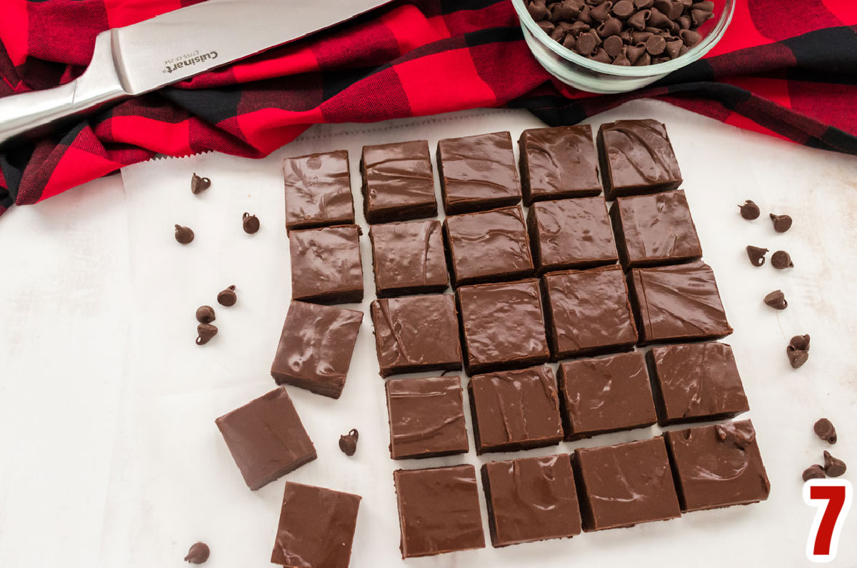 Overhead shot of 24 pieces of Easy Chocolate Fudge arranged in rows on a piece of parchment paper with a silver knife, buffalo check table linen and a ramekin filled with chocolate chips.