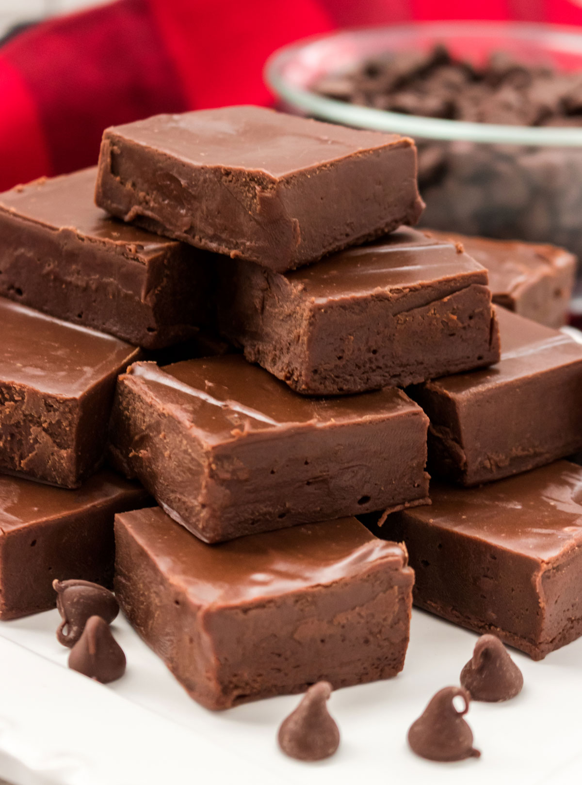 Closeup on a stack of Easy Chocolate Fudge sitting on a white surface in front of a red linen and a glass bowl filled with chocolate chips.