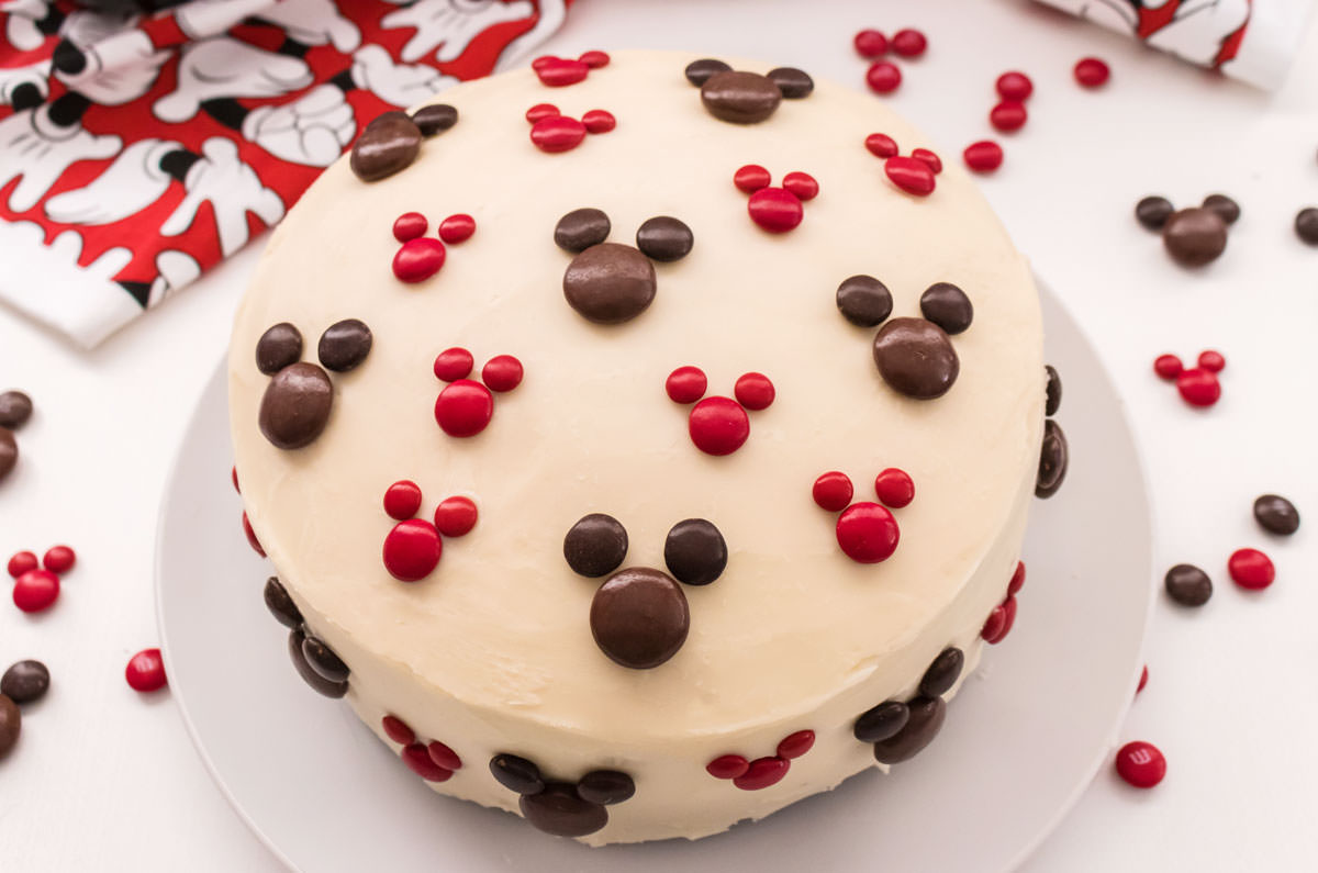 Overhead view of an Easy Mickey Mouse Cake sitting on a white cake plate on a white surface surrounded by M&M's and a Mickey Mouse kitchen towel.