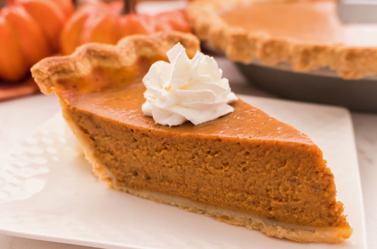 Closeup on a piece of Homemade Pumpkin Pie sitting on a white dessert plate in front of decorative pumpkins.