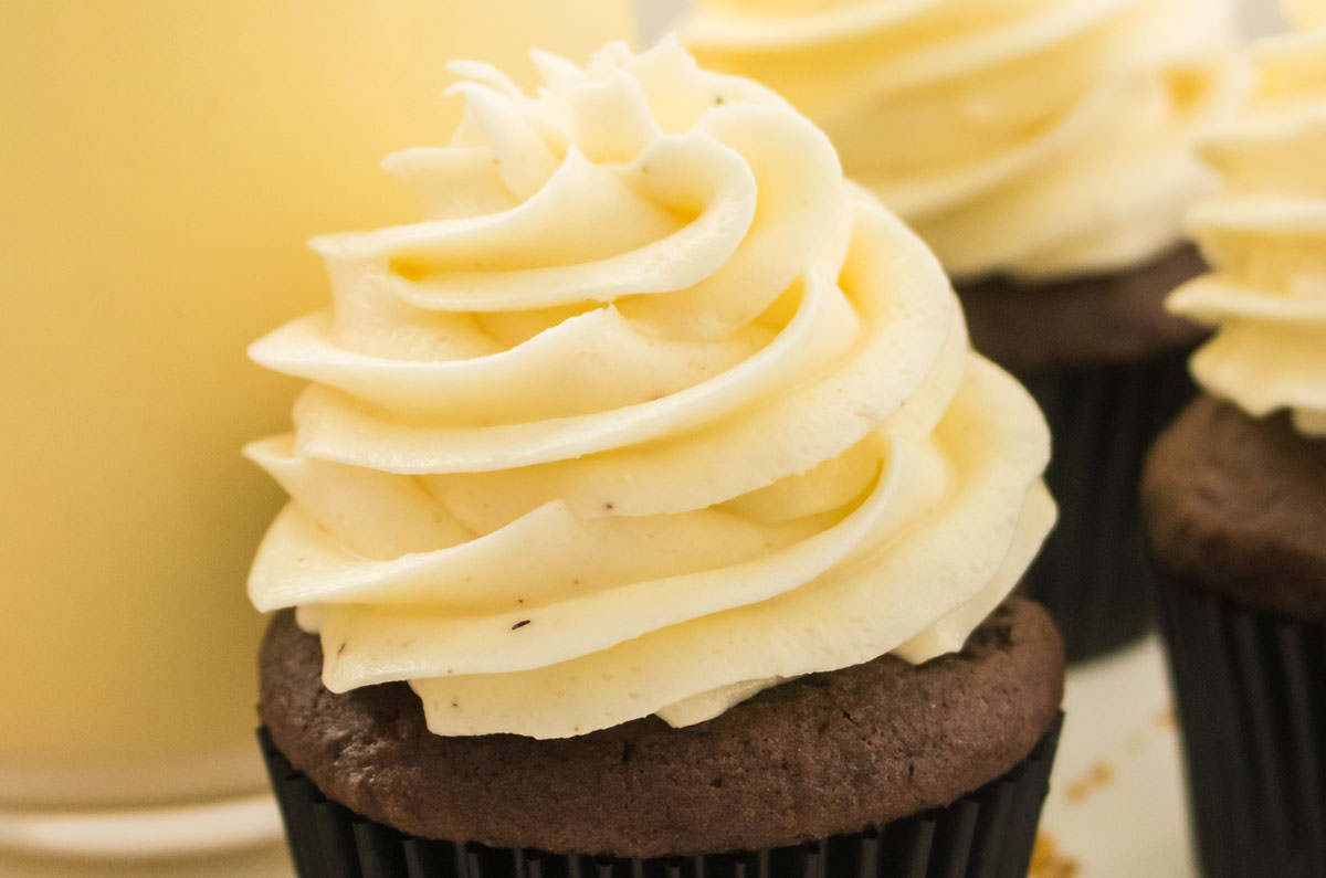Closeup on a cupcake topped with The Best Eggnog Buttercream Frosting sitting on a white surface in front of a glass of Eggnog.