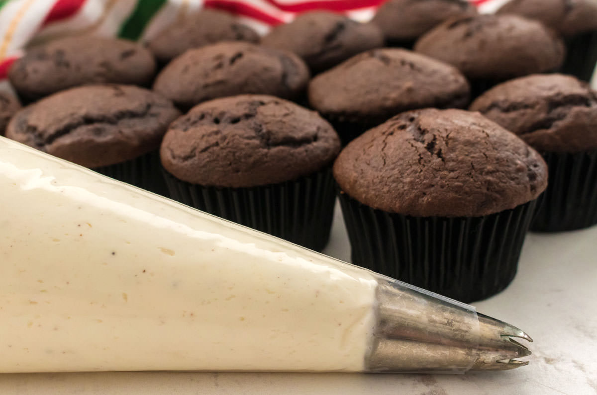 Closeup on a a decorating bag filled with Eggnog Frosting sitting on a white table in front of a batch of unfrosted cupcakes.