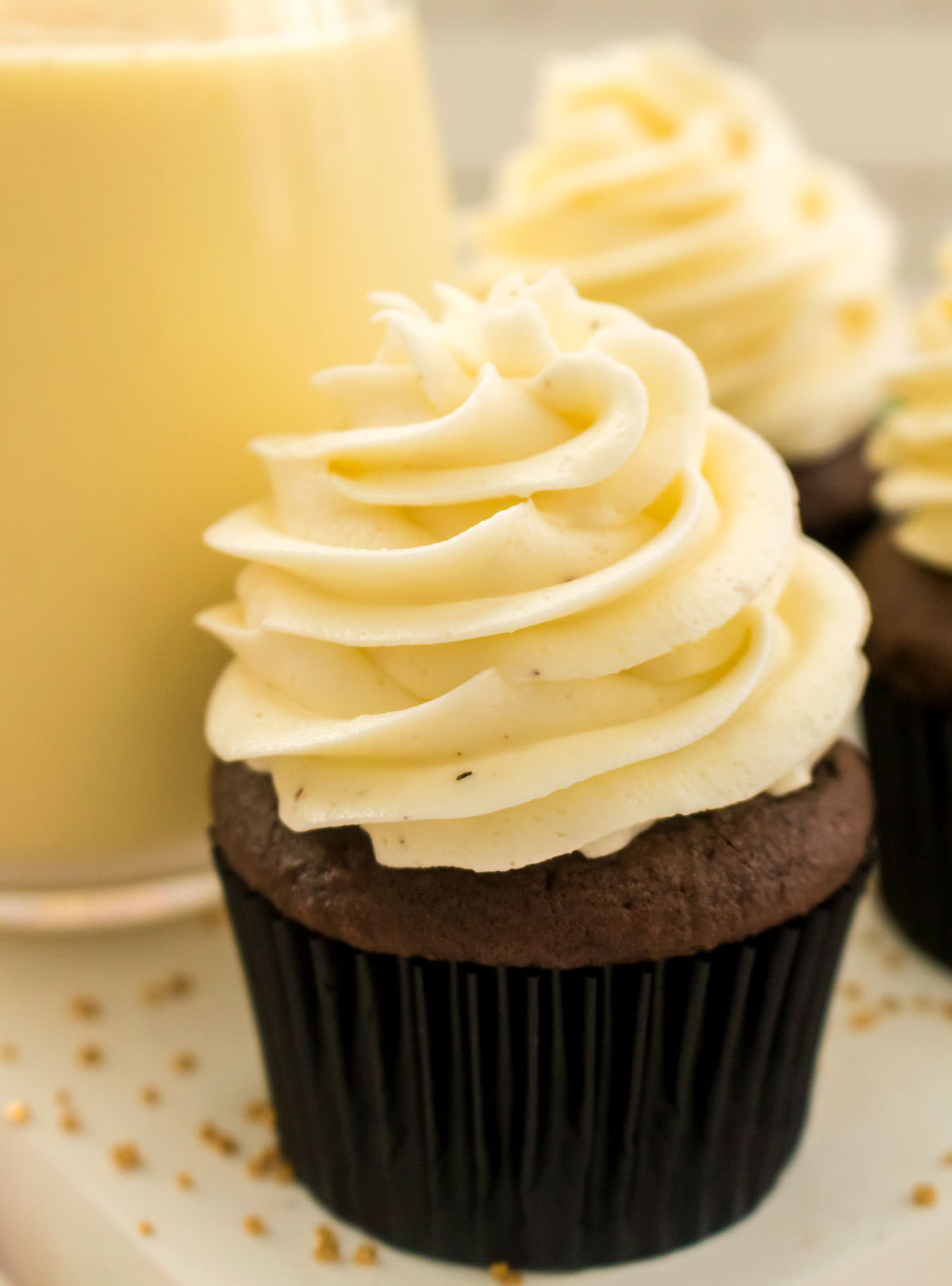 Closeup of a chocolate cupcake topped with The Best Eggnog Buttercream Frosting sitting on a white table in front of a glass of Eggnog.