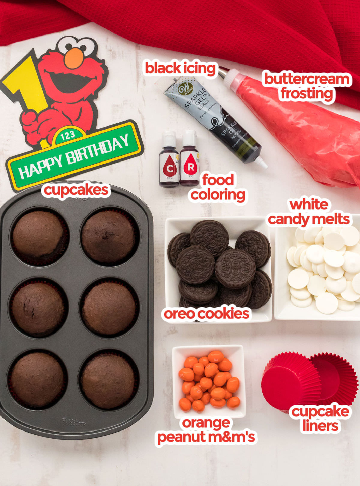 All the ingredients you will need to make Elmo Cupcakes including cupcakes, buttercream frosting, black icing, food coloring, candy melts, oreo cookies, orange peanut m&m's and red cupcake liners.