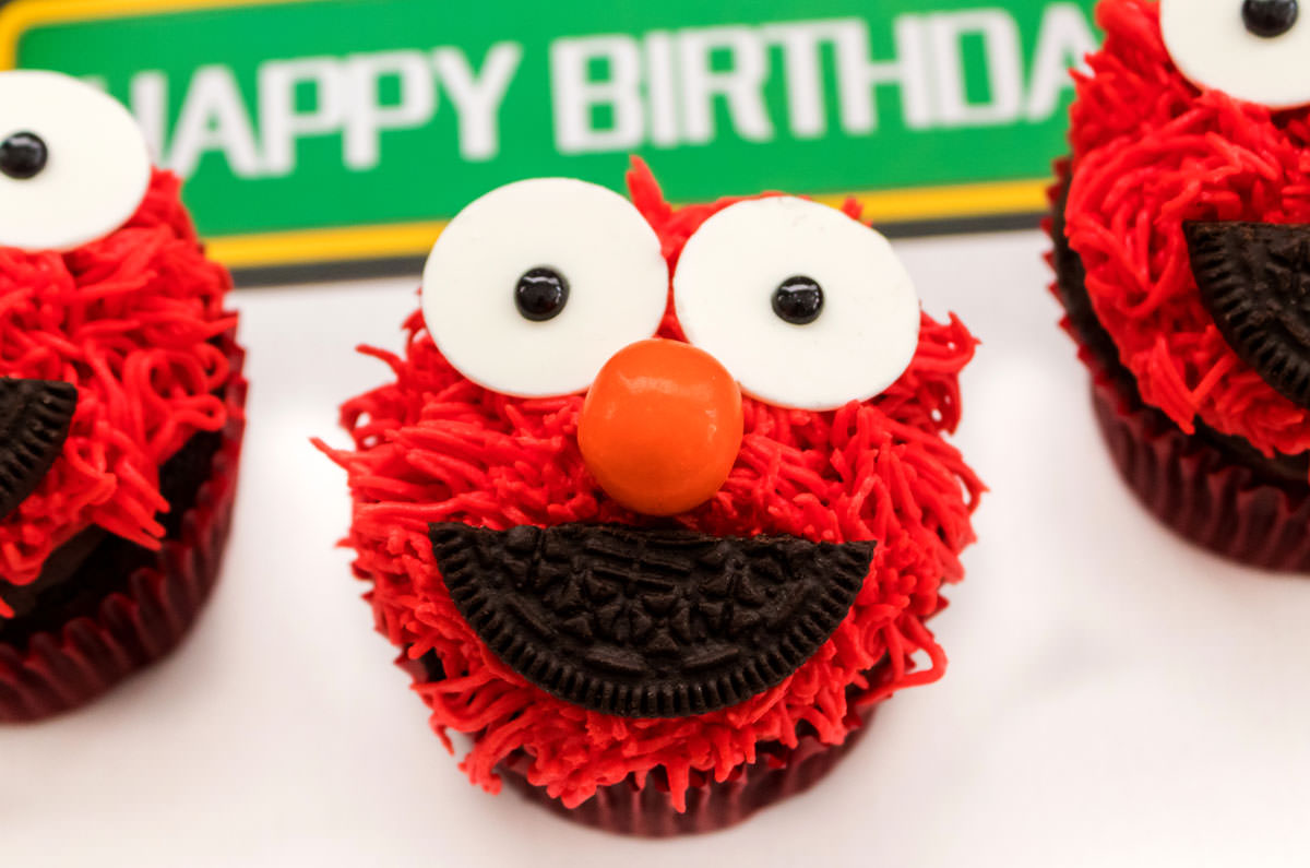 Close on three Elmo Cupcakes sitting on a white surface in front of a Happy Birthday Sesame Street Sign.