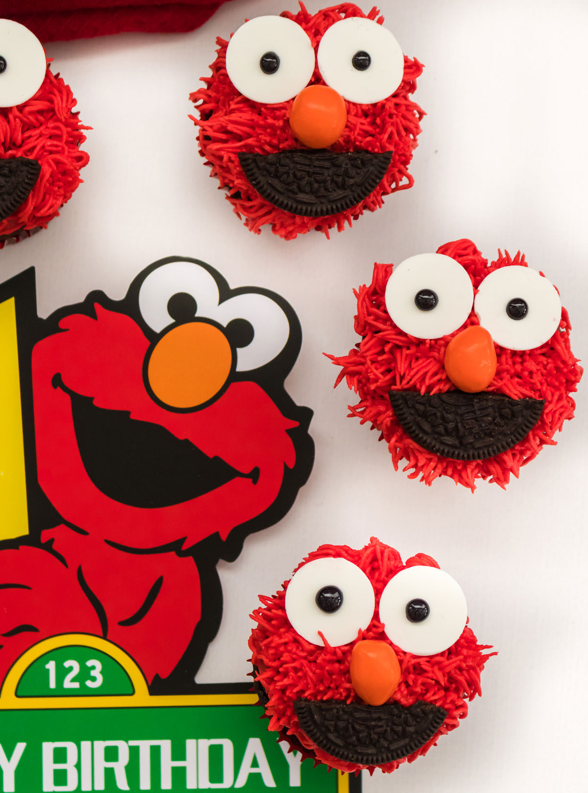 Closeup on three Elmo Cupcakes sitting on a white surface next to a Elmo Happy Birthday Banner.