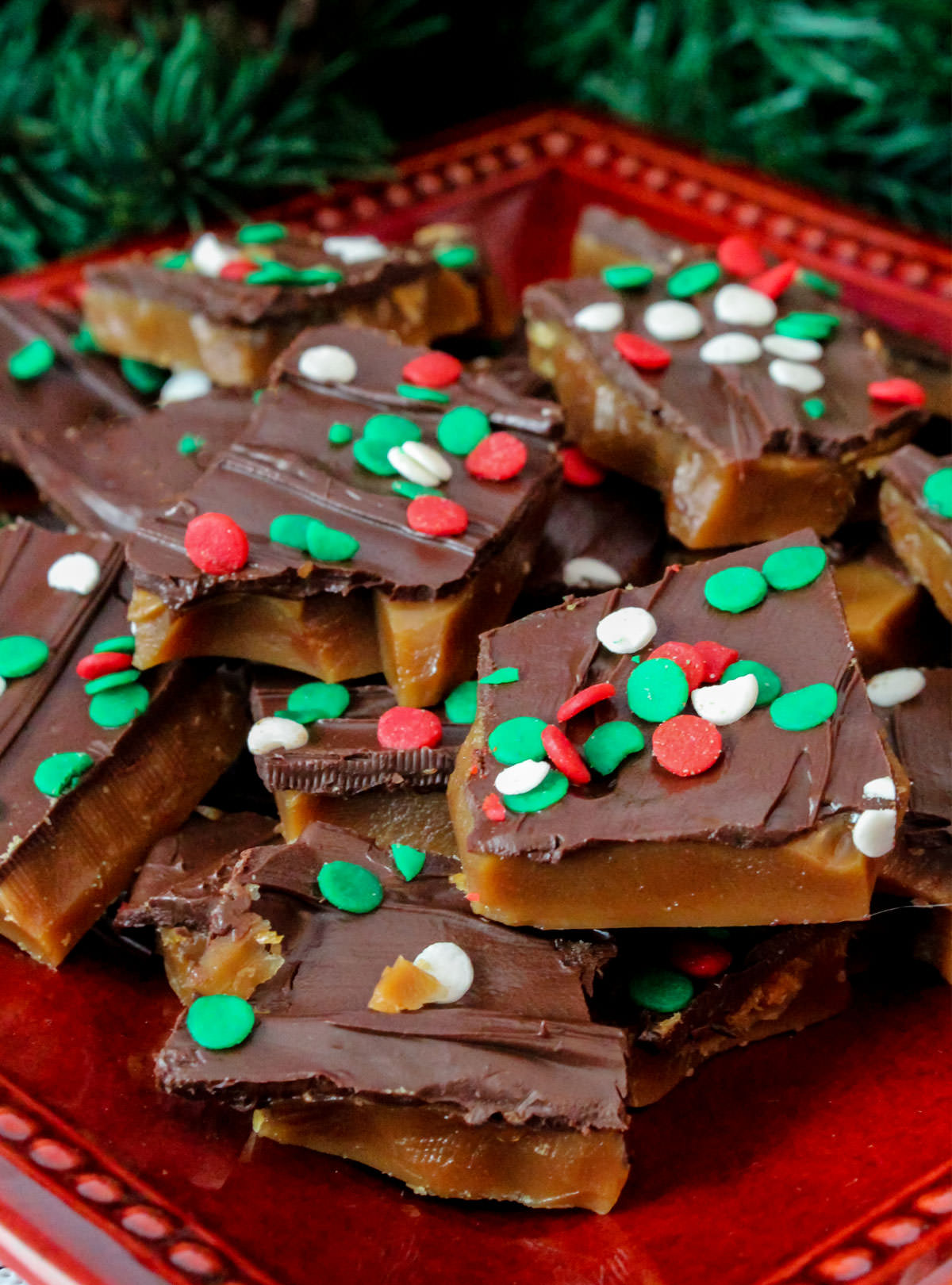 Closeup on a batch of Homemade English Toffee sitting on a red plate surrounded by Christmas decorations.