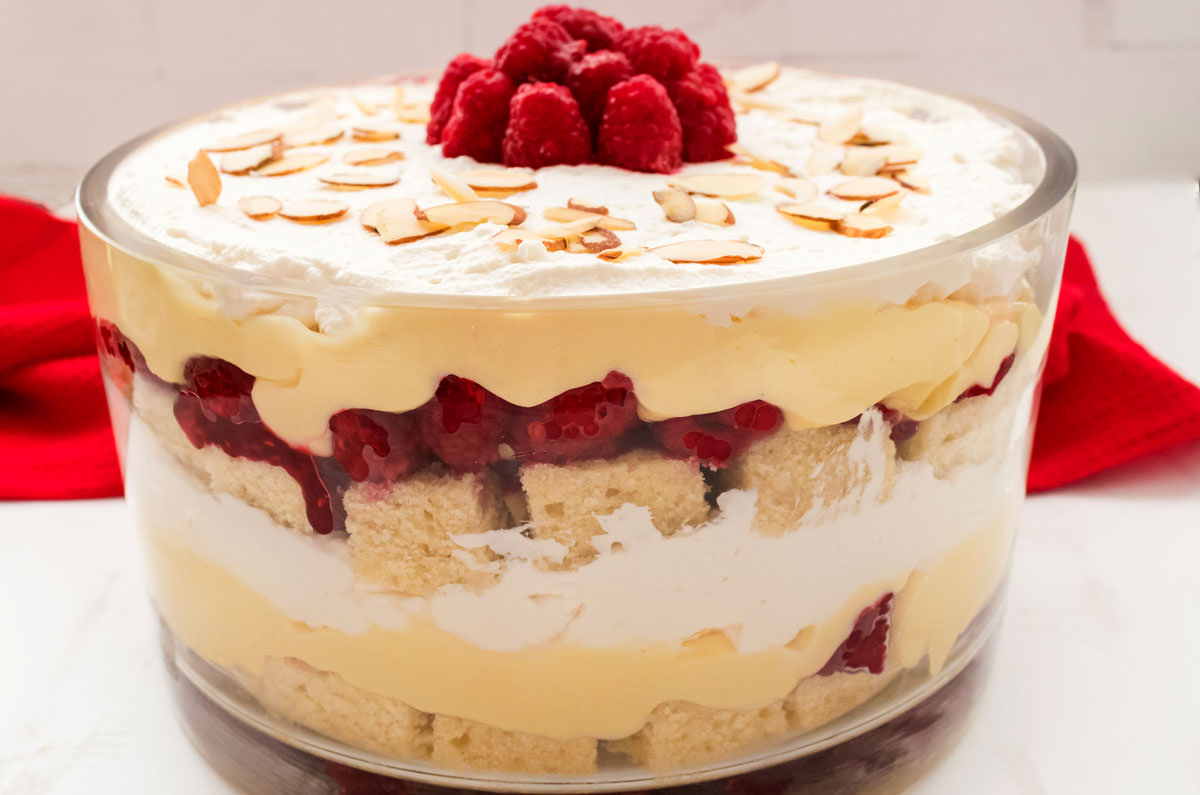 A large glass bowl filled with English Trifle sitting on a white surface in front of a red kitchen towel.