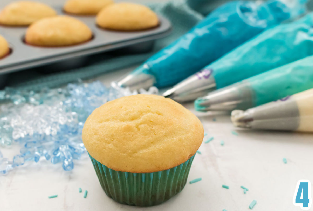 Closeup on a vanilla cupcake sitting in front of four decorator bags filled with frosting and a tin full of vanilla cupcakes.