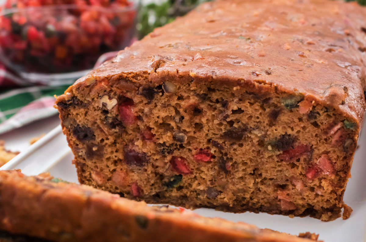 Closeup on a loaf of Fruit Cake sitting on a white platter with Christmas Decorations in the background.