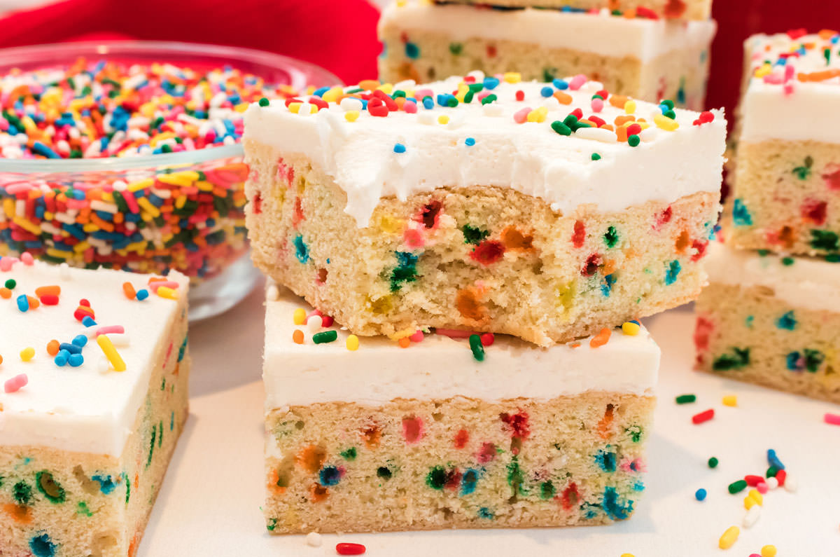 Closeup on a stack of two Funfetti Sugar Cookie Bars, one with a bite taken out of it, sitting surrounded by other cookie bars and a glass bowl filled with rainbow sprinkles.