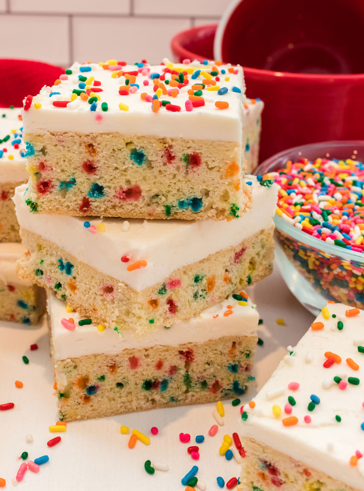 Closeup on a stack of three Funfetti Sugar Cookie Bars sitting next to a glass bowl full of rainbow sprinkles on a white table.