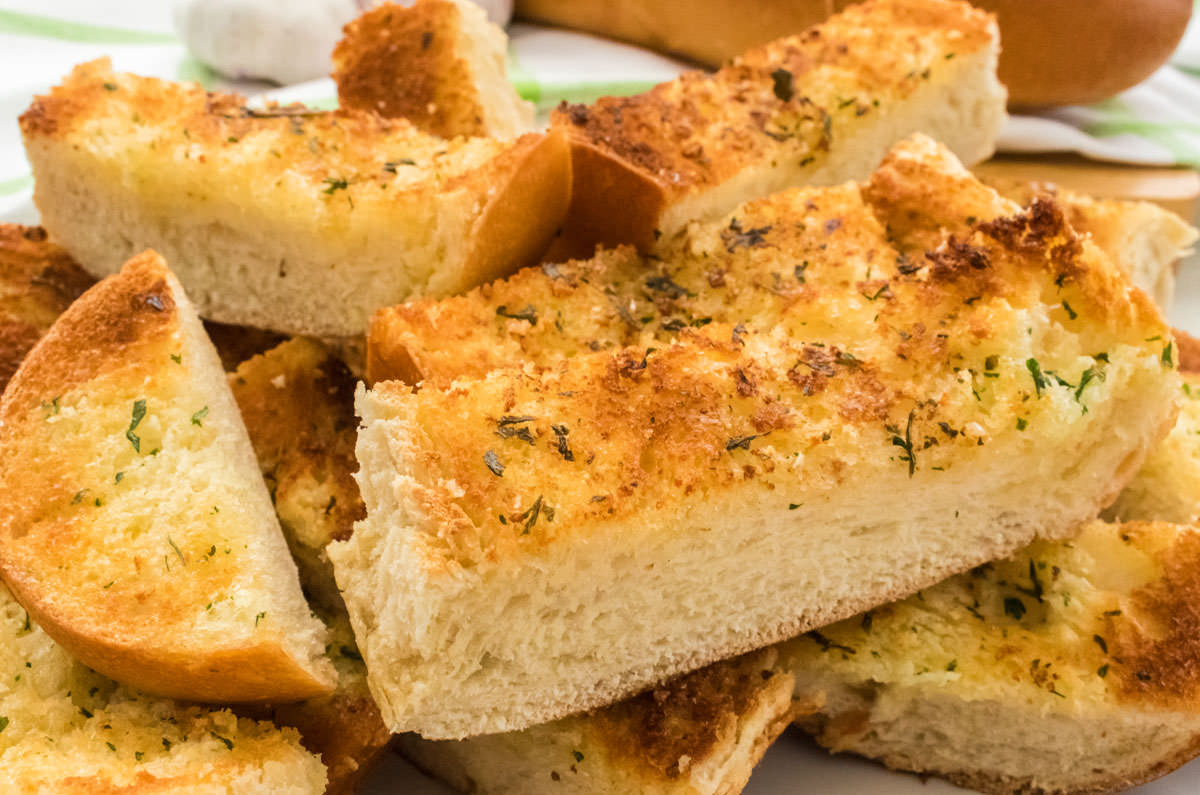 Closeup on a large stack of Homemade Garlic Bread pieces sitting on a white plate in front of a loaf of French Bread and Garlic.