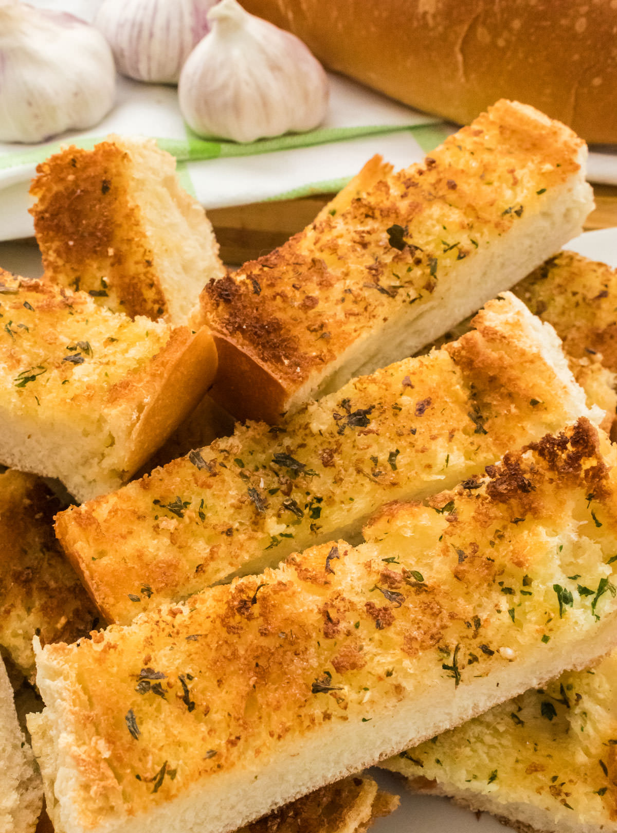 Closeup on a stack of Garlic Bread pieces stacked on a white serving dish on a table in front of heads of garlic and a loaf of French Bread.