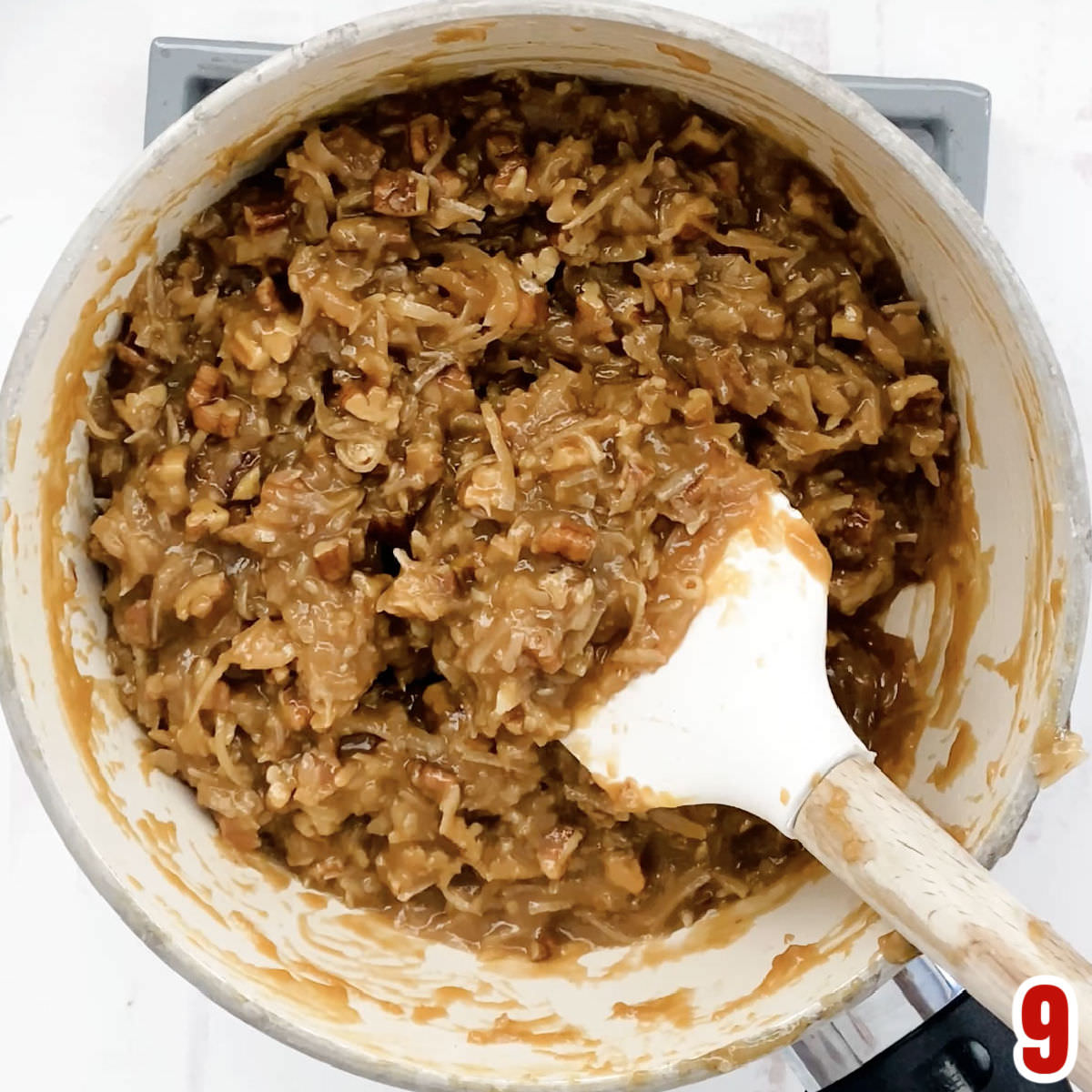 Overhead shot of a white saucepan filled with cooling German Chocolate Frosting and a white wooden spatula.