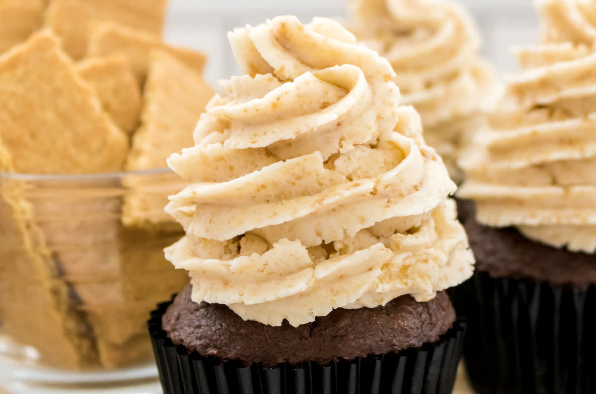 Closeup on three cupcakes topped with Graham Cracker Buttercream Frosting sitting next to a bowl filled with Graham Crackers.