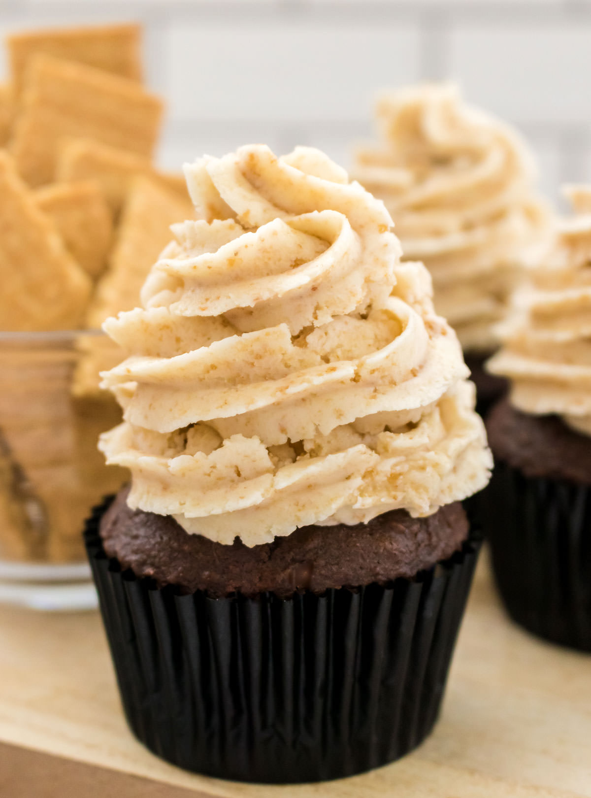 Closeup on a chocolate cupcake topped with Graham Cracker Buttercream Frosting sitting in front of a glass bowl filled with Graham Crackers.
