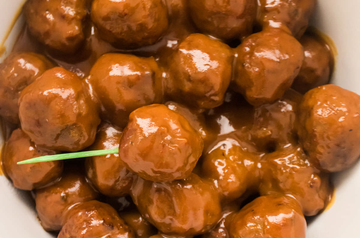 Closeup of a top of a white Crockpot filled with Grape Jelly Meatballs, a single meatball has a toothpick in it.