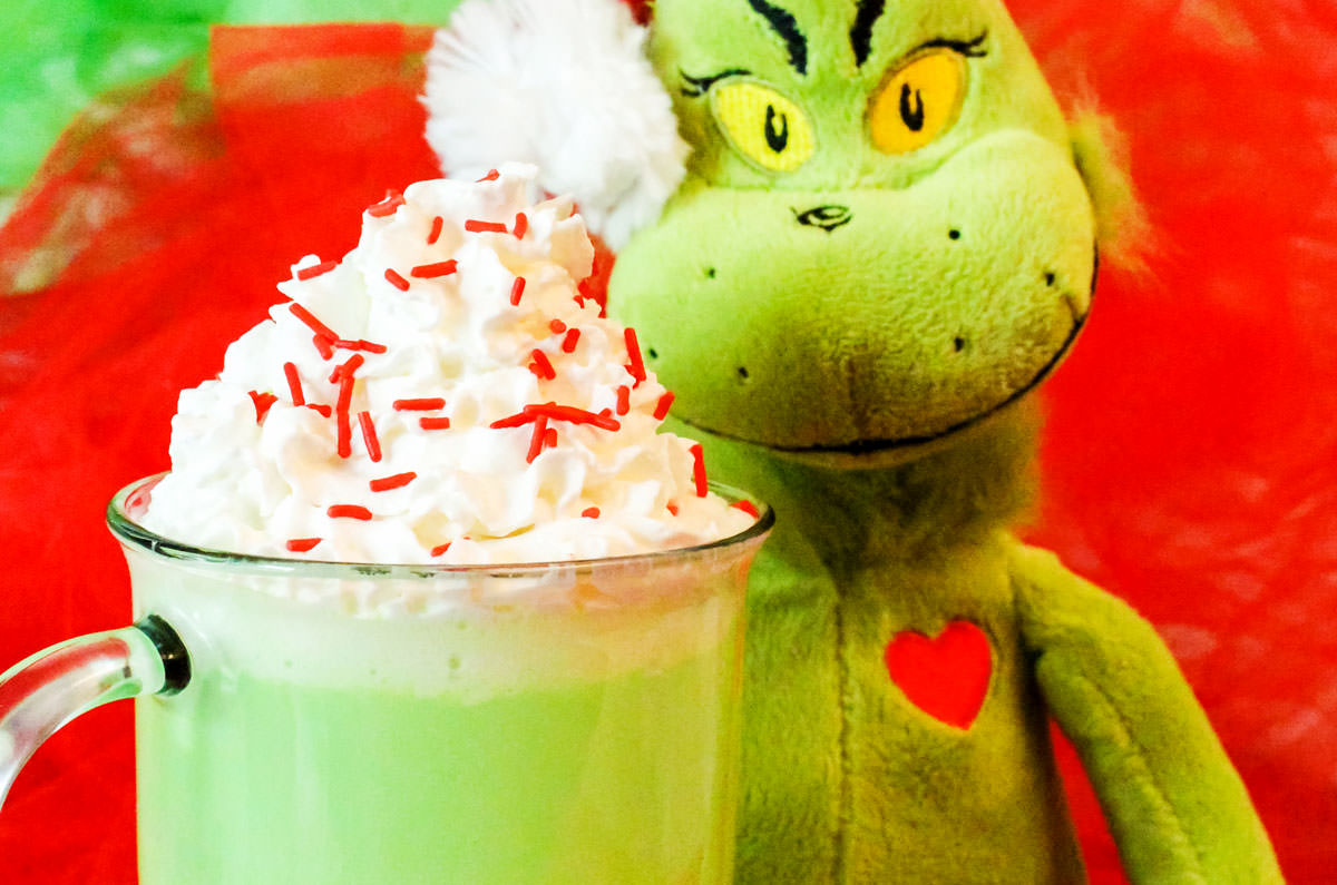 Glass mug filled with Grinch Hot Vanilla Milk topped with Whipped Cream and Red Sprinkles sitting next to a Grinch stuffed animal.