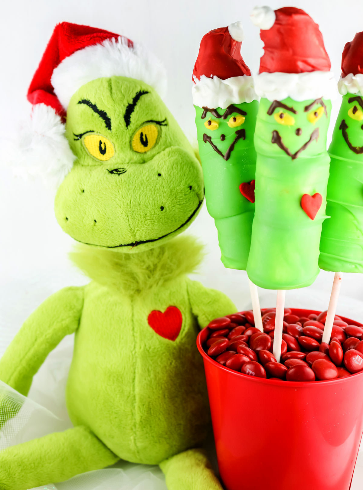 Closeup on a Grinch doll sitting next to a red pail holding three Grinch Marshmallow Pops.