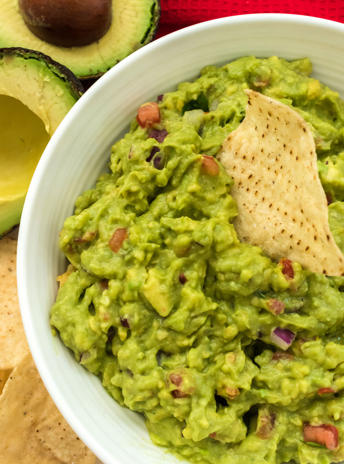 Closeup on a white bowl filled with Guacamole with tortilla chips and avocados surrounding the bowl.