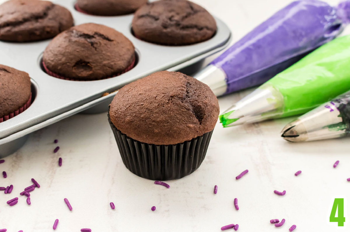 Chocolate Cupcake sitting on a white table surrounded by a cupcake pan filled with cupcakes and decorating bags filled with purple, green and black frosting.