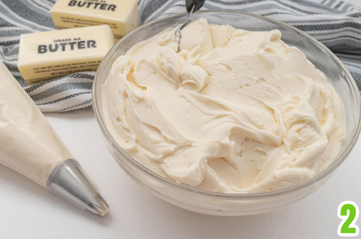 Clear glass bowl filled with buttercream frosting sitting next to butter, a decorating bag filled with frosting and a black and white towel.