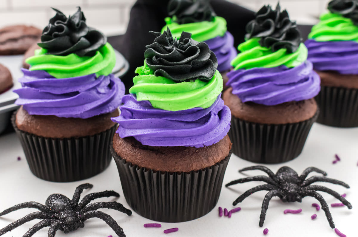 Closeup on five Bewitched Halloween Cupcakes sitting on a white table surrounded by toy spiders, black towel and a cupcake pan filled with chocolate cupcakes.