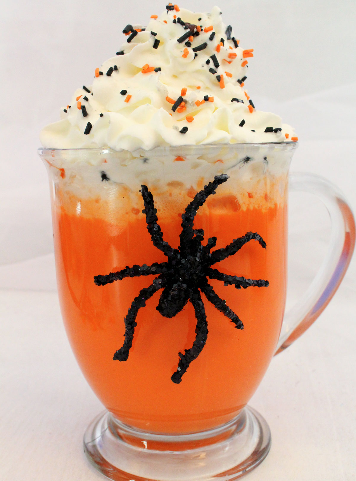 Closeup on a glass of Halloween Hot Vanilla Milk sitting on a white table with a toy spider on the glass.