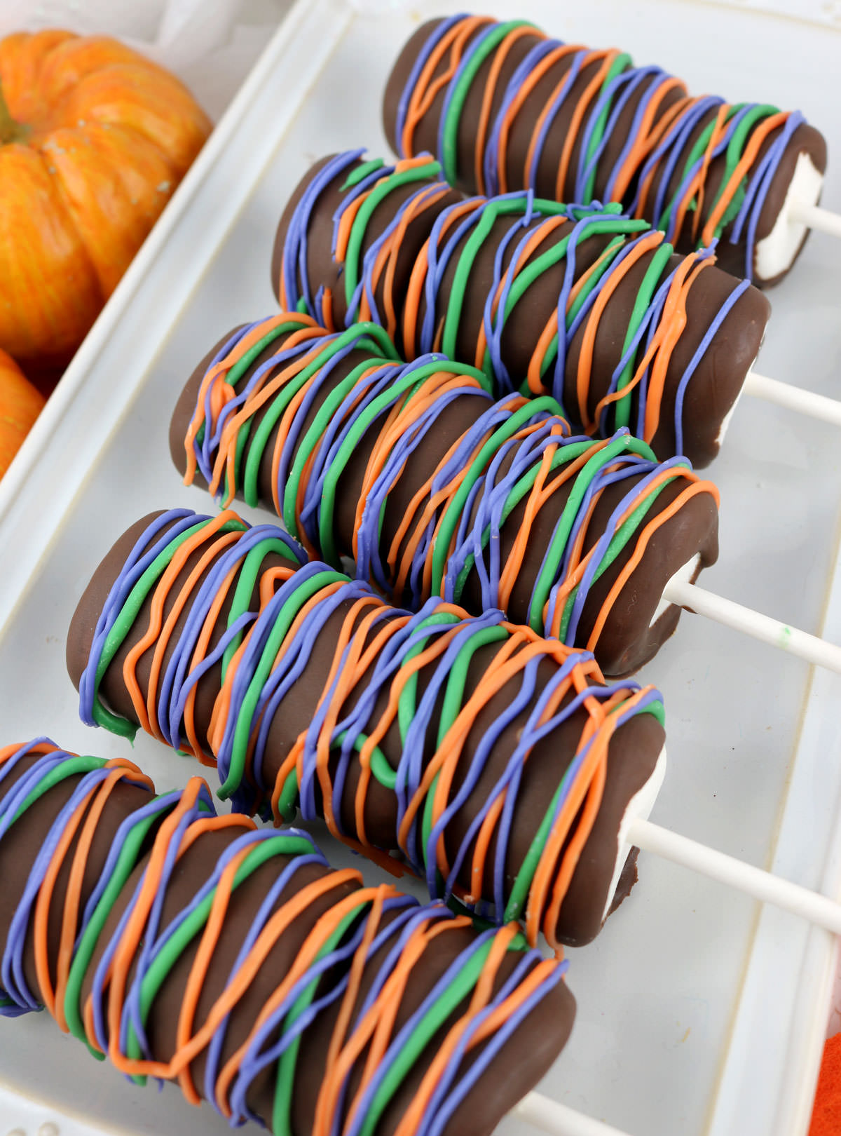 Closeup of five Halloween Marshmallow Pops laying on a white serving platter with decorative pumpkins off to the left.