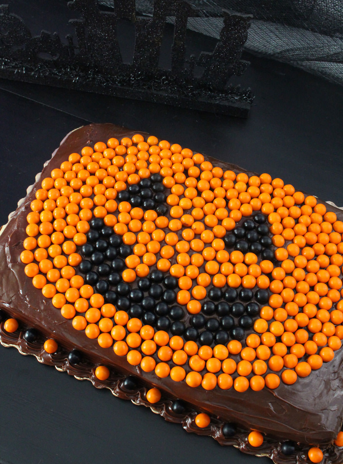 Closeup on a Chocolate Sheet Cake decorated with a Pumpkin made from black and orange Sixlets sitting on a black tabletop.