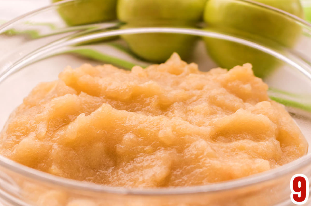 Closeup on a glass bowl filled with The Best Homemade Applesauce sitting in front of three green apples.