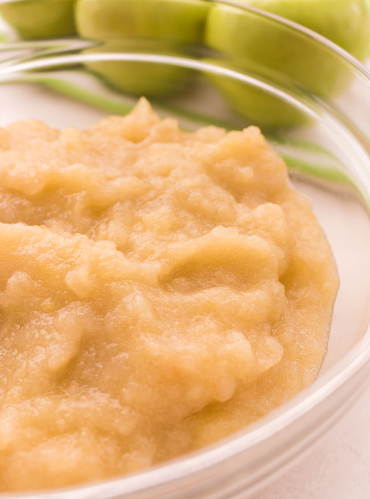 Closeup on a glass bowl filled with The Best Homemade Applesauce sitting on a white table in front of green apples.