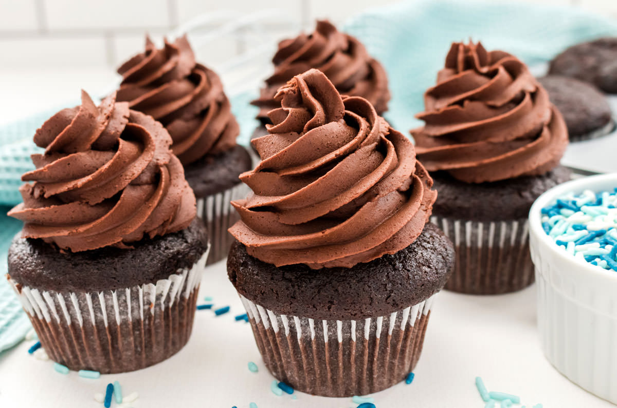 Close up on five frosted Homemade Chocolate Cupcakes sitting on a white table surrounded by sprinkles and a blue table linen.