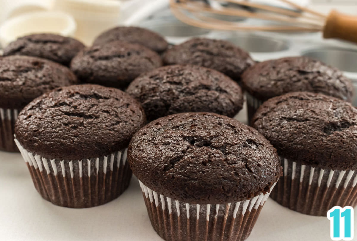 Closeup on a batch of chocolate cupcakes sitting on a white table in front of a cupcake tin and a whisk.