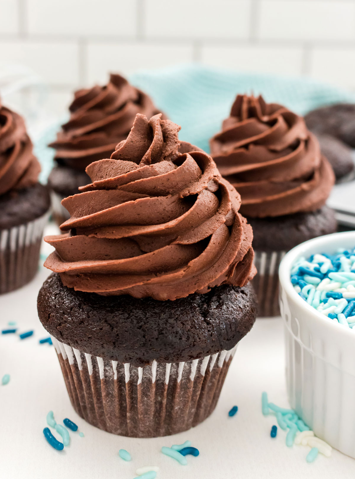 Close up on four Homemade Chocolate Cupcakes sitting on a white table surrounded by sprinkles and a blue table linen.