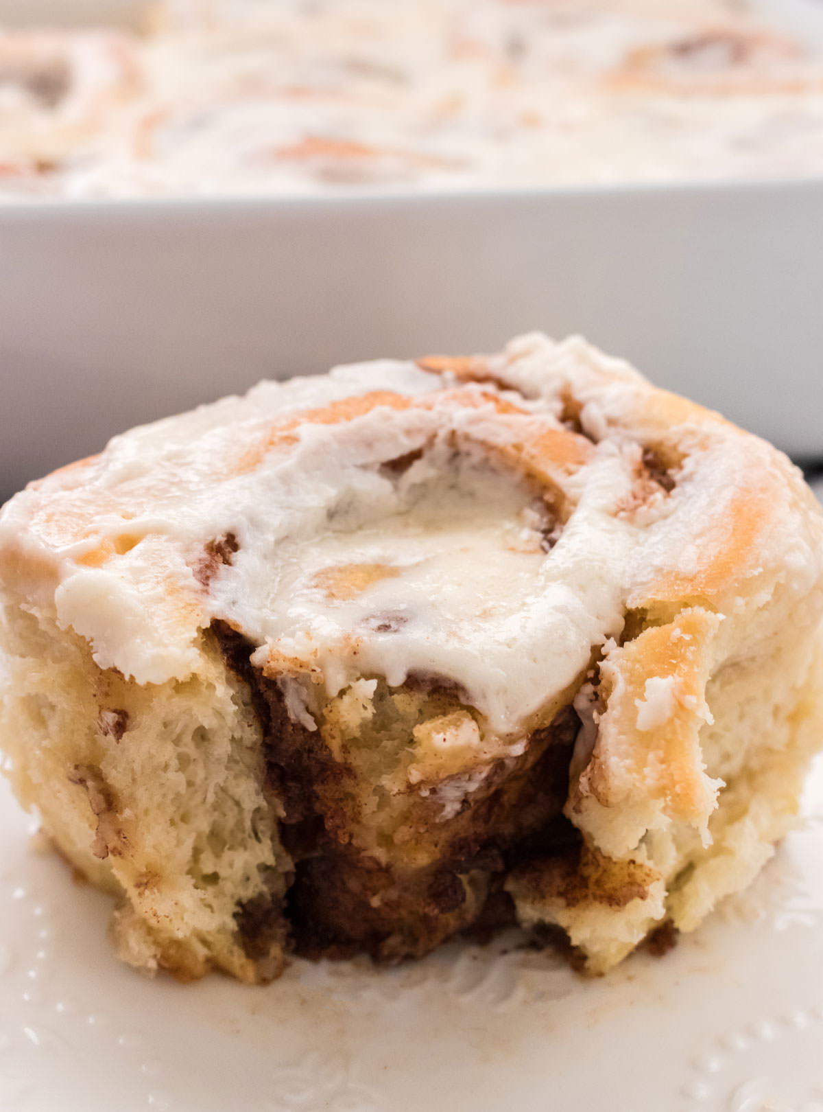 Homemade Cinnamon Roll with a bite taken out of it sitting on a white plate in front of a pan of Cinnamon rolls.