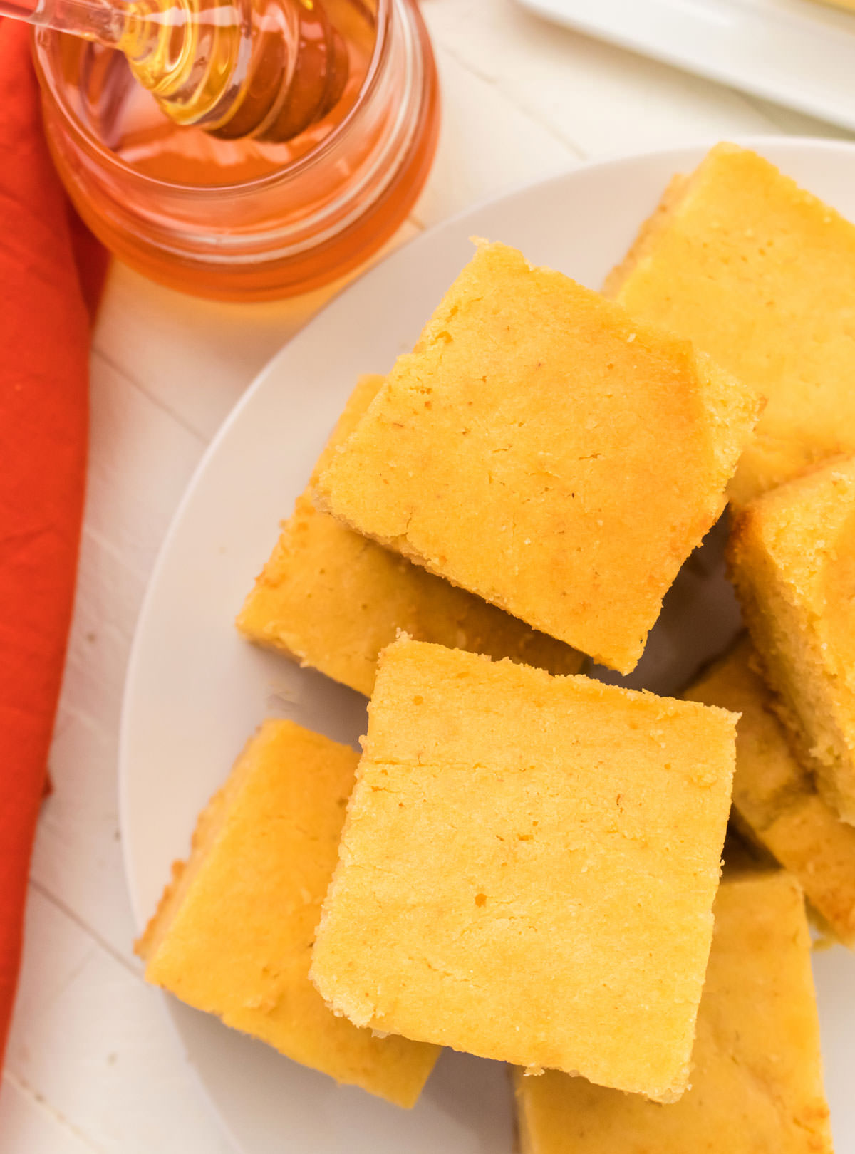 Closeup on eight pieces of Homemade Cornbread sitting on a white serving plate which is sitting on a white table next to a orange table linen and a jar of honey.